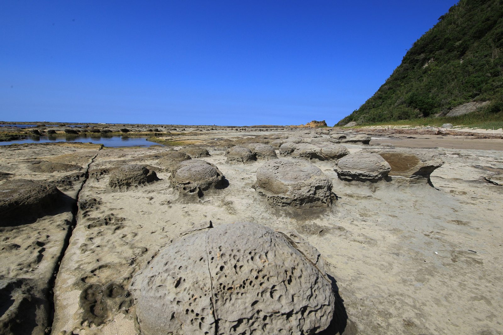Flat rocky shore platform with rounded rock masses scattered across it under a clear blue sky, with a green hillside at right
