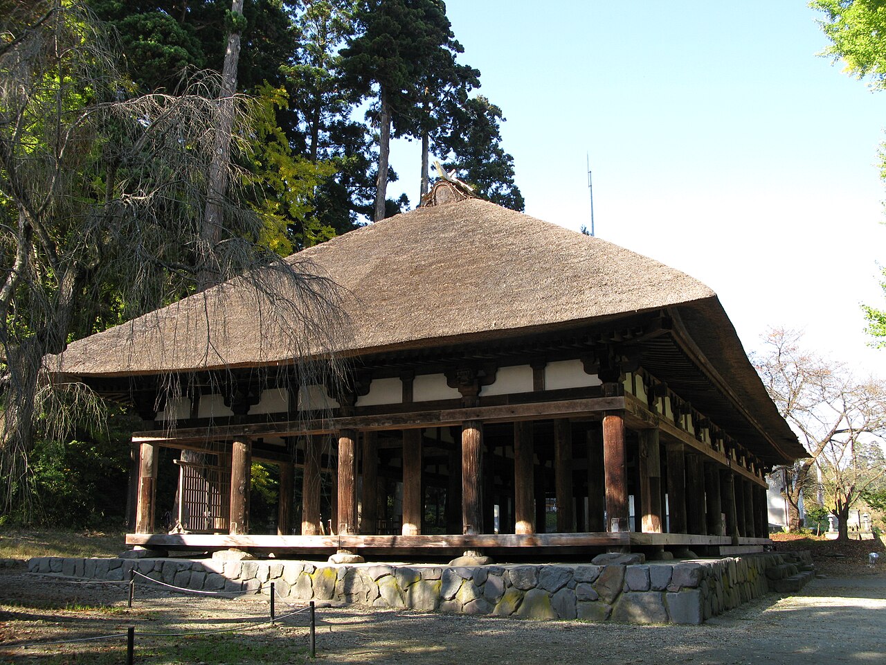 A long wooden hall with no side walls, rows of round pillars on a stone base and a sharply pitched thatched roof, set against tall cedars under a blue sky