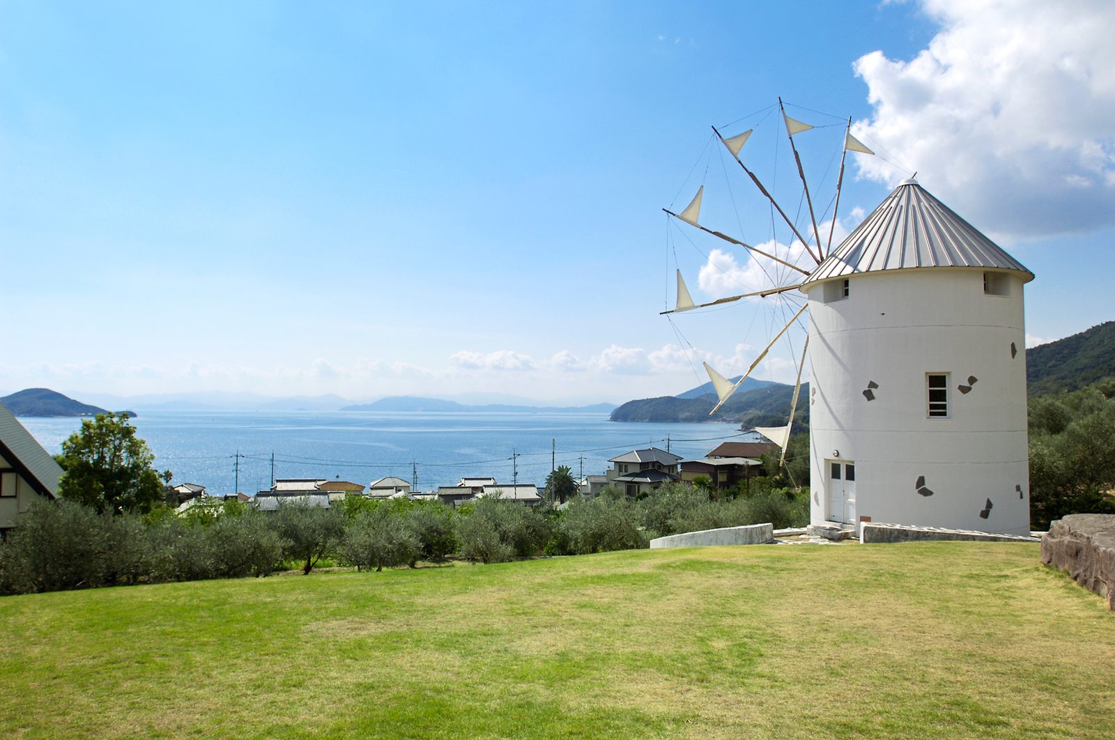 A white windmill on a lawn at Shodoshima Olive Park, with olive trees, a row of coastal houses and the Seto Inland Sea beyond.