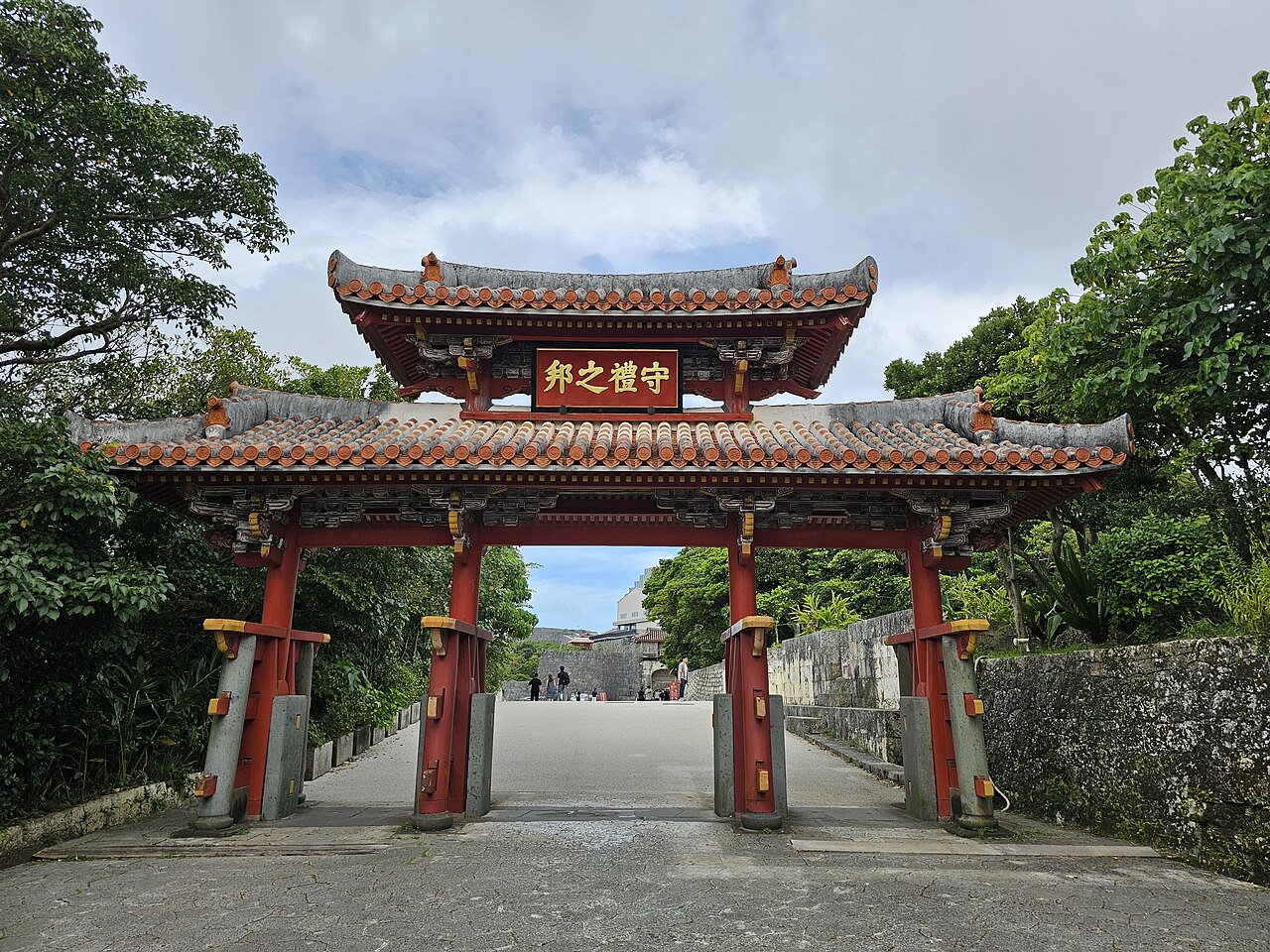 Shurei-mon, a red two-roof gate at Shuri Castle with a gold-lettered plaque reading 守禮之邦, seen from the stone path leading up to it and flanked by trees, with a few people visible in the distance beyond the gate.