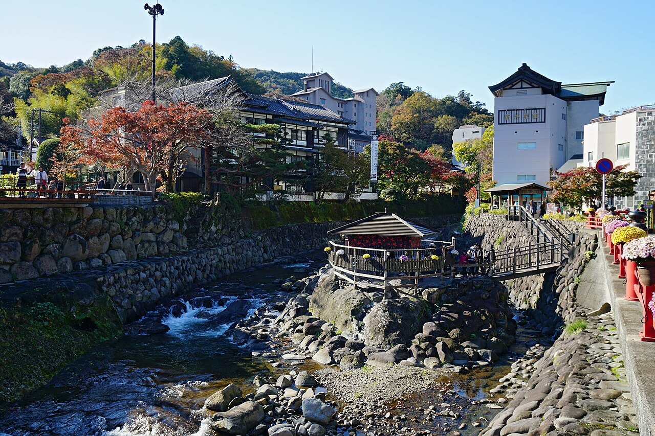 The shallow, rocky bed of the Katsura River at Shuzenji, with a small roofed pavilion standing on a rock midstream, stone walls on both banks and wooden inns and autumn foliage behind.