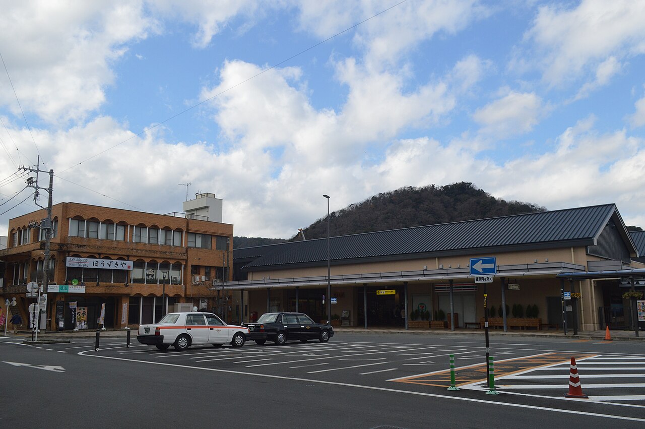 The forecourt of Shuzenji Station under a grey sky, with two taxis waiting in front of the low, dark-roofed station building and a wooded hillside close behind.