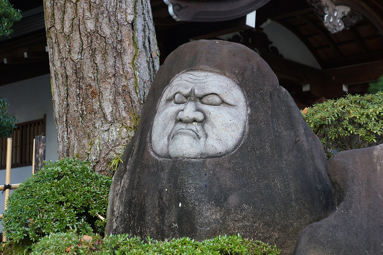 A large dark boulder in the grounds of Shuzenji's temple, its rounded face picked out in white like a hooded head, with a pine trunk, clipped shrubs and the eaves of the temple behind.