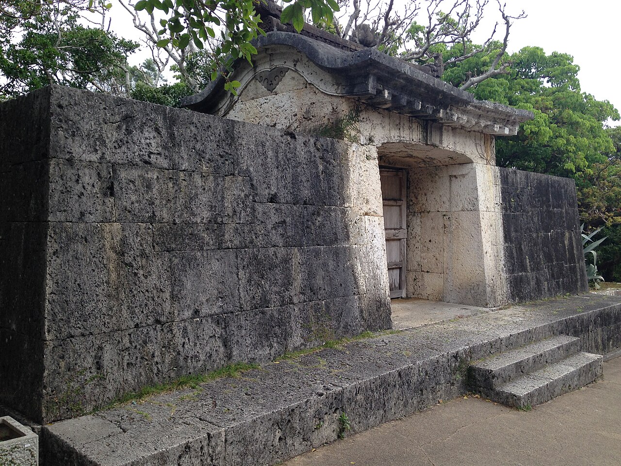 The stone gate of Sonohyan-utaki, a coral-limestone archway with a carved, upturned tile-roof lintel, set into a low stone wall near Shuri Castle, with trees behind it.