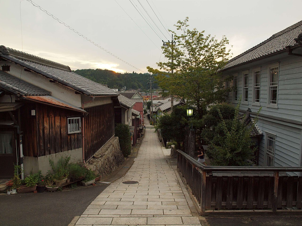 A stone-paved lane sloping down between old houses in Akechi at dusk, with a lit street lamp