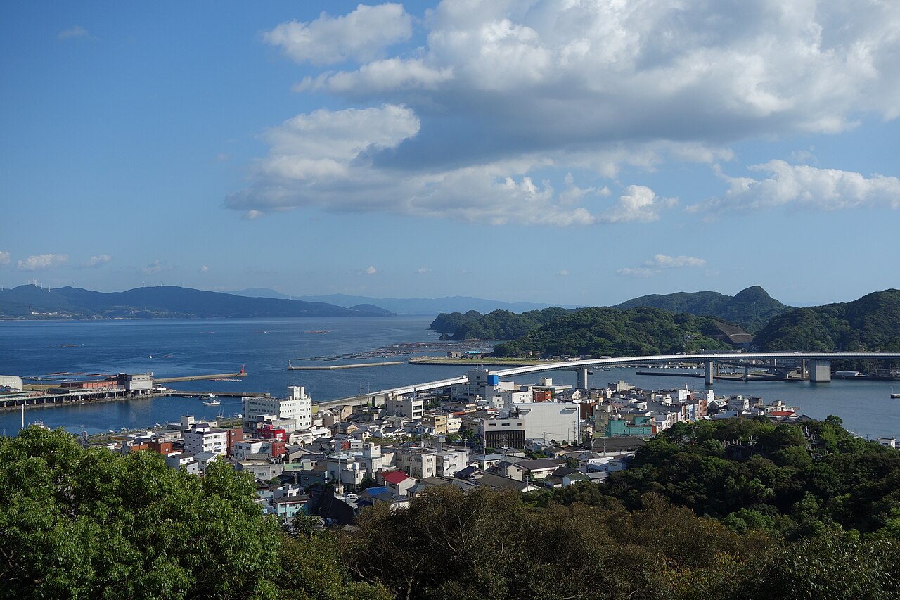 A small harbor town seen from a hilltop park in clear weather, with white buildings, fishing piers, a long curving bridge and green headlands over a blue bay
