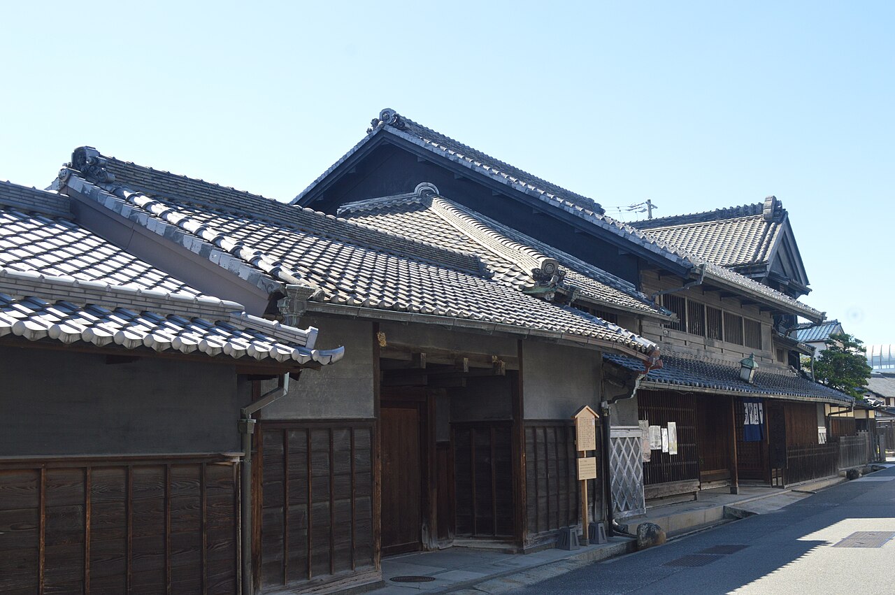 Old tile-roofed merchant houses with dark wooden walls and a small signboard along a sunlit street in Arimatsu under a clear sky.