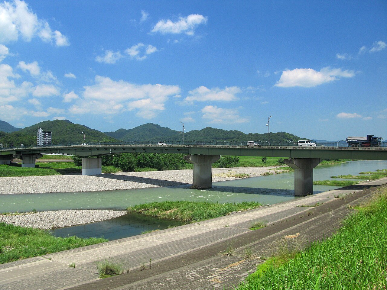The Watarase River at Ashikaga with clear green water over gravel bars, a low concrete road bridge and forested hills under a blue sky