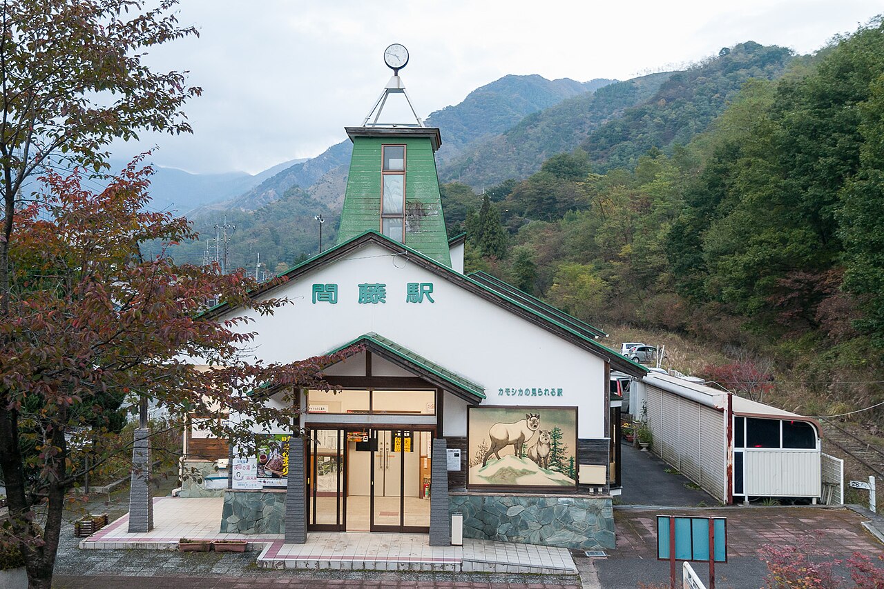 A white and green Matō Station building with a small clock tower stands in a mountain valley under cloud, with wooded slopes and autumn-coloured trees around it