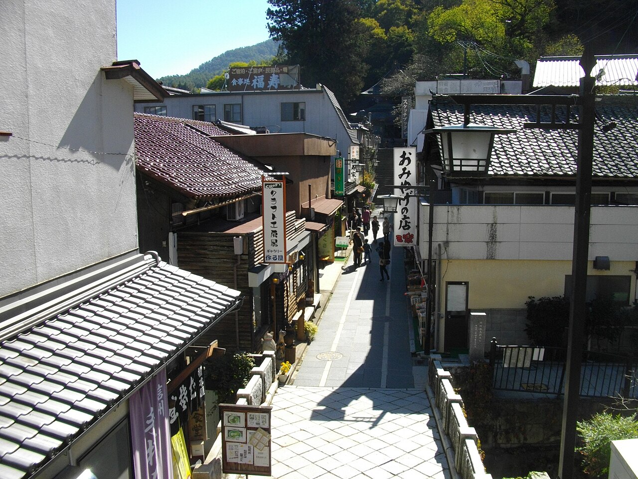 A narrow paved lane lined with tiled-roof shops and souvenir signs in a hot-spring town, with a few visitors walking and forested hills beyond.