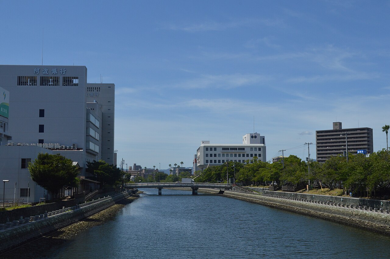 A wide calm river lined with stone embankments and trees running through the city of Tokushima under a blue sky, with a low bridge in the distance