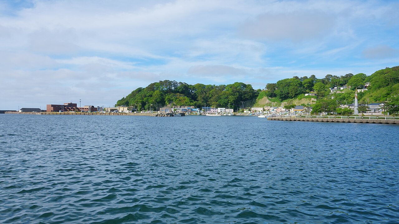 A calm blue harbor in Fukaura with a small fishing town and green wooded hills along the far shore under a partly cloudy sky.