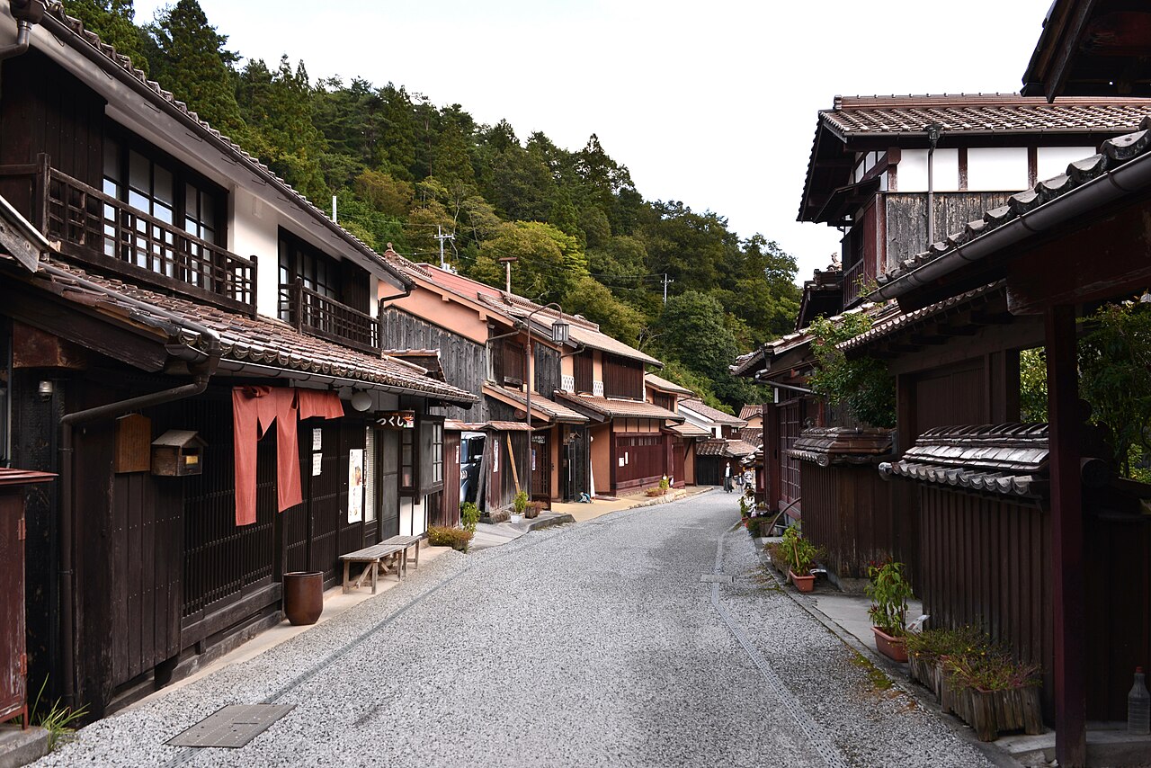 A wide, quiet gravel lane in Fukiya between old lattice-fronted houses, with wooded hills rising behind the rooftops