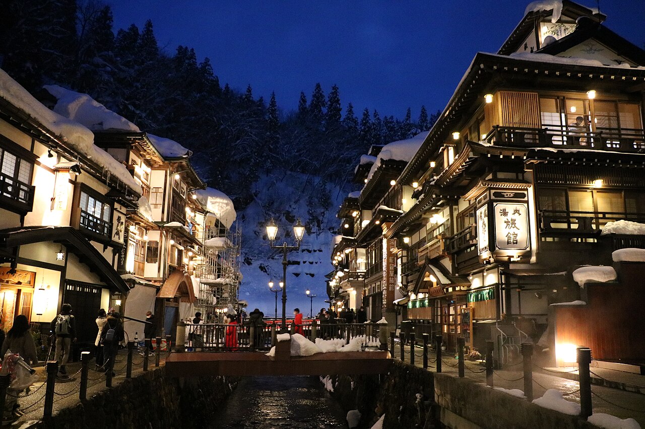 A snowy dusk street in an onsen town with lit wooden inns on both sides and a small bridge over a stream.