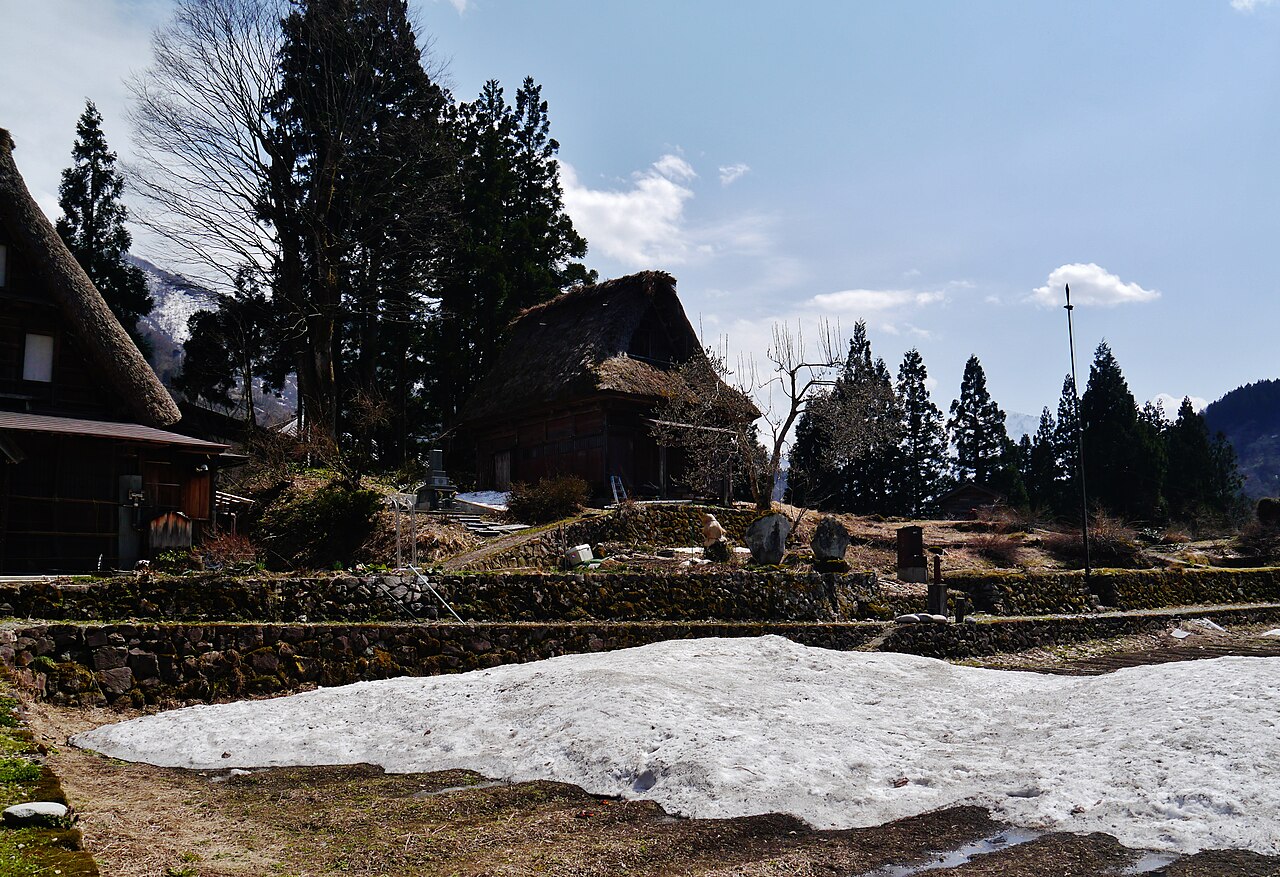Thatched gassho-zukuri houses above stone terrace walls in Ainokura, with late snow lying on the field in front