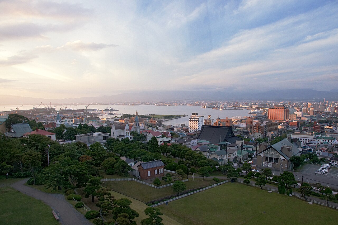 Elevated view over the Motomachi district of Hakodate at dusk, with a lawn and a small red-brick building among trees in the foreground, rooftops and church spires in the middle, and the bay and distant mountains under a pale cloudy sky