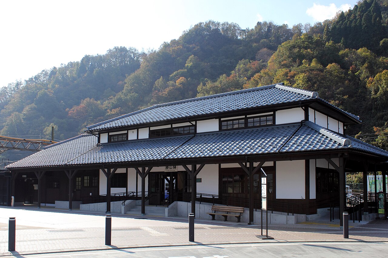 A dark-tiled wooden station building with a sweeping roof stands before forested mountains under a pale sky