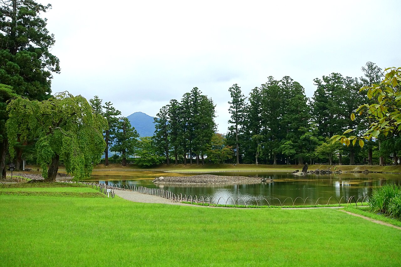 A green lawn in the foreground sloping to a pond with a small gravel island, tall conifers along the far bank and a rounded mountain behind under a cloudy sky.