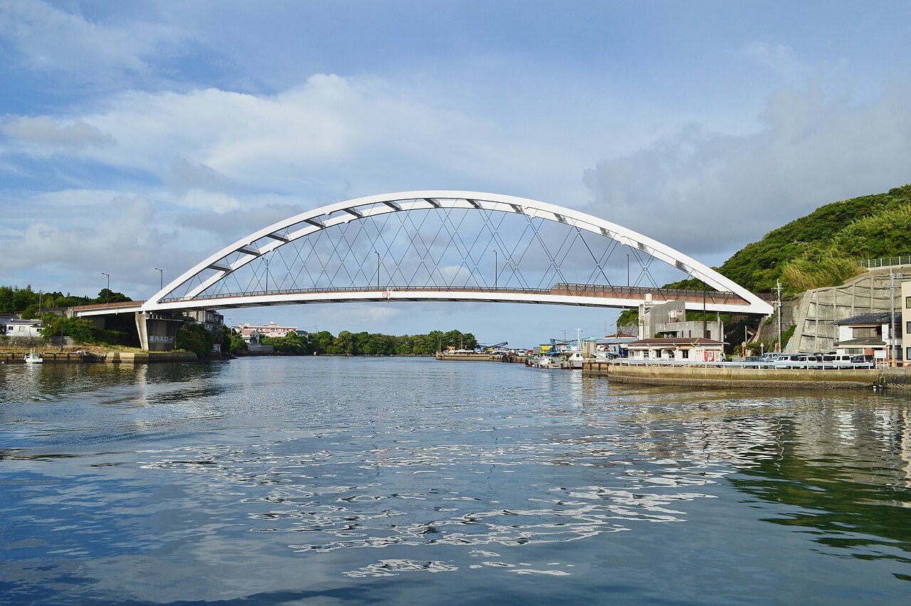 A white arched bridge spanning a calm harbor channel in Gonoura, Iki Island, with small boats and waterfront buildings beside green hills.