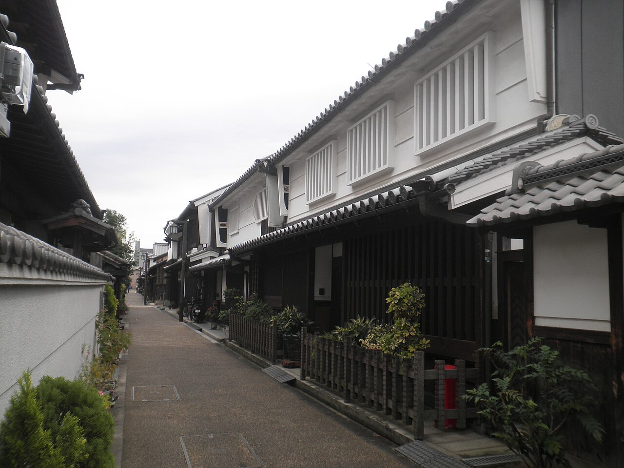 A quiet lane in Imai-cho with two-storey wooden merchant houses, white plaster walls, and dark timber lattice fronts, potted plants along the pavement, no people in view.