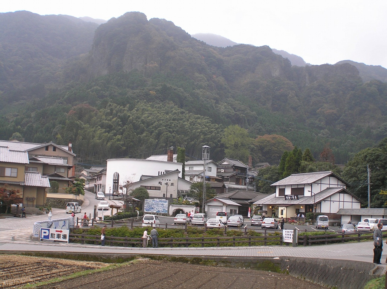 A small kiln village in a green valley, with white-walled workshops and a car park below steep forested rock cliffs under overcast sky
