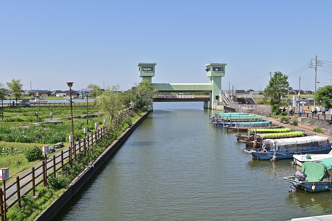 A straight green waterway runs toward a pale-green floodgate tower under a clear blue sky, with iris beds and a wooden fence on the left and moored pleasure boats on the right