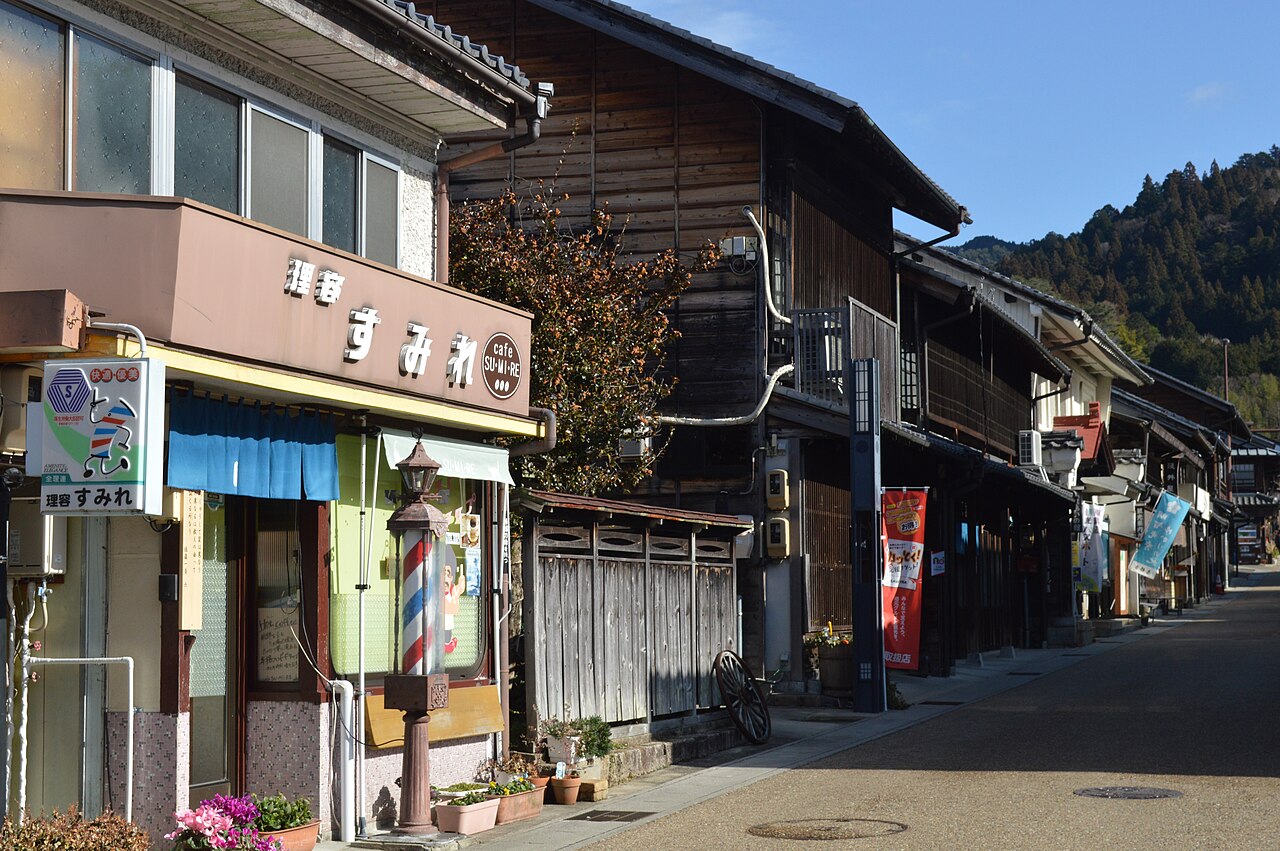 A sunlit street of old wooden townhouses with a barber pole and shop signs at left, and forested hills rising behind the rooftops.