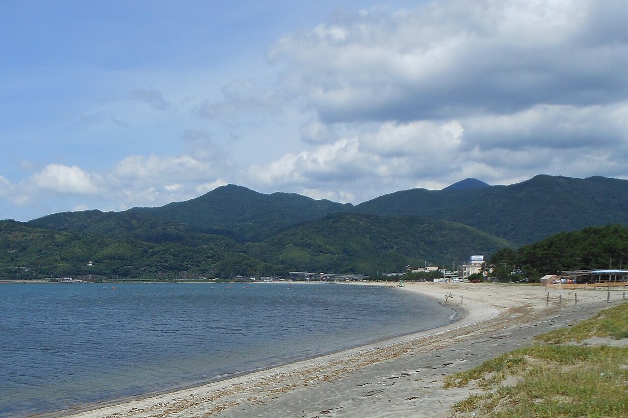 The curve of Hamasaki beach on Karatsu Bay at low tide, with forested hills behind the shoreline.