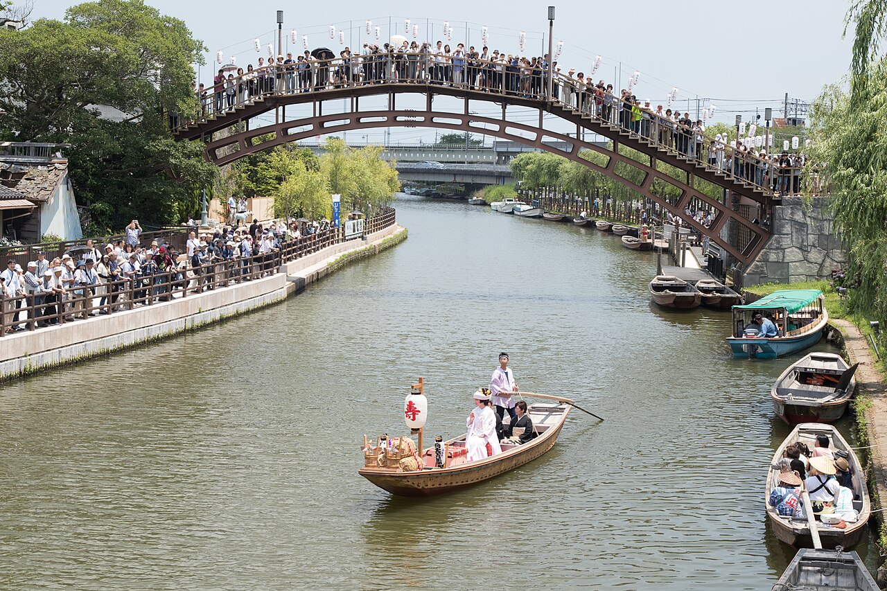 A bridal boat gliding along the willow-lined canal at Itako with an arched wooden bridge and onlookers behind