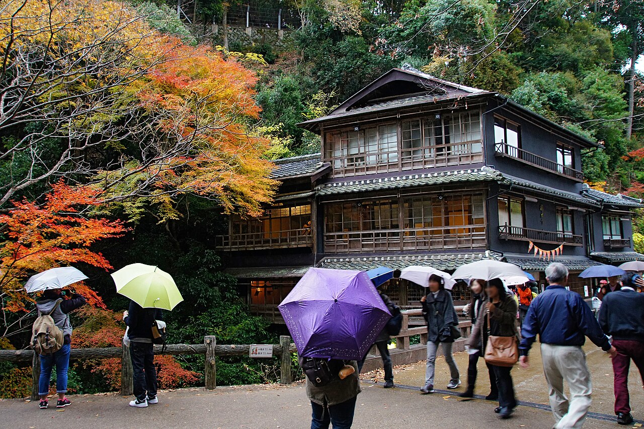 A dark-timbered multi-story wooden building among autumn maple trees in Minoh, with visitors carrying umbrellas passing in front