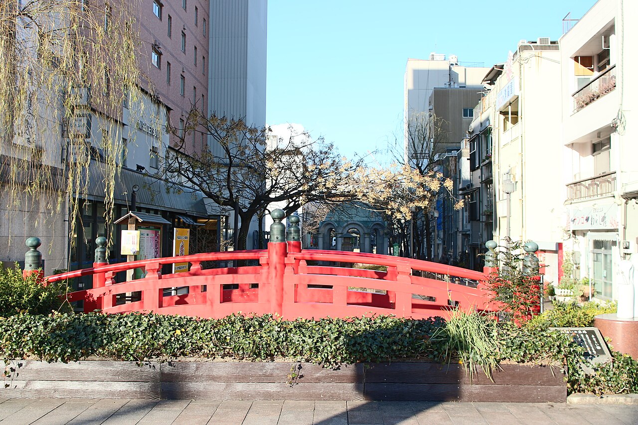 A red arched bridge with green post caps among city buildings in central Kochi, with a weeping willow beside it and a blue sky above