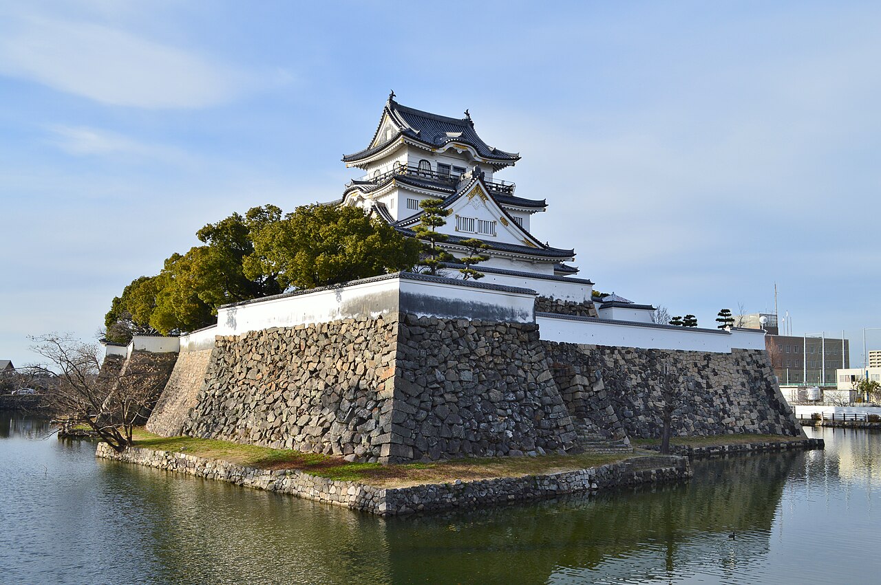 Kishiwada Castle's keep standing on a stone-walled corner of the moat, with pine trees on the embankment and calm water reflecting a pale evening sky.