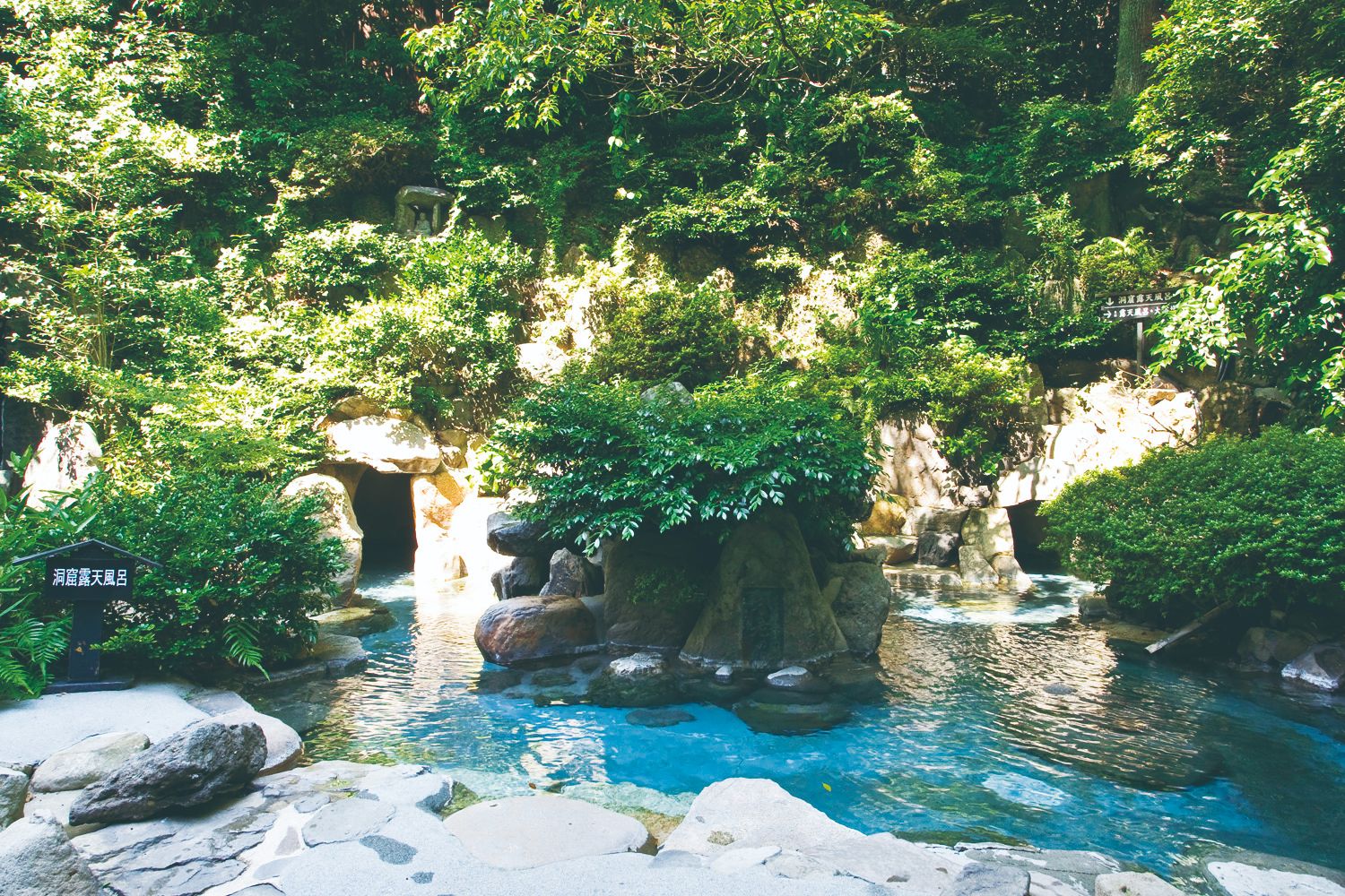 A rocky open-air hot spring bath of turquoise water in front of a cave-like grotto, surrounded by dense green trees and ferns, with a small signboard at the left.