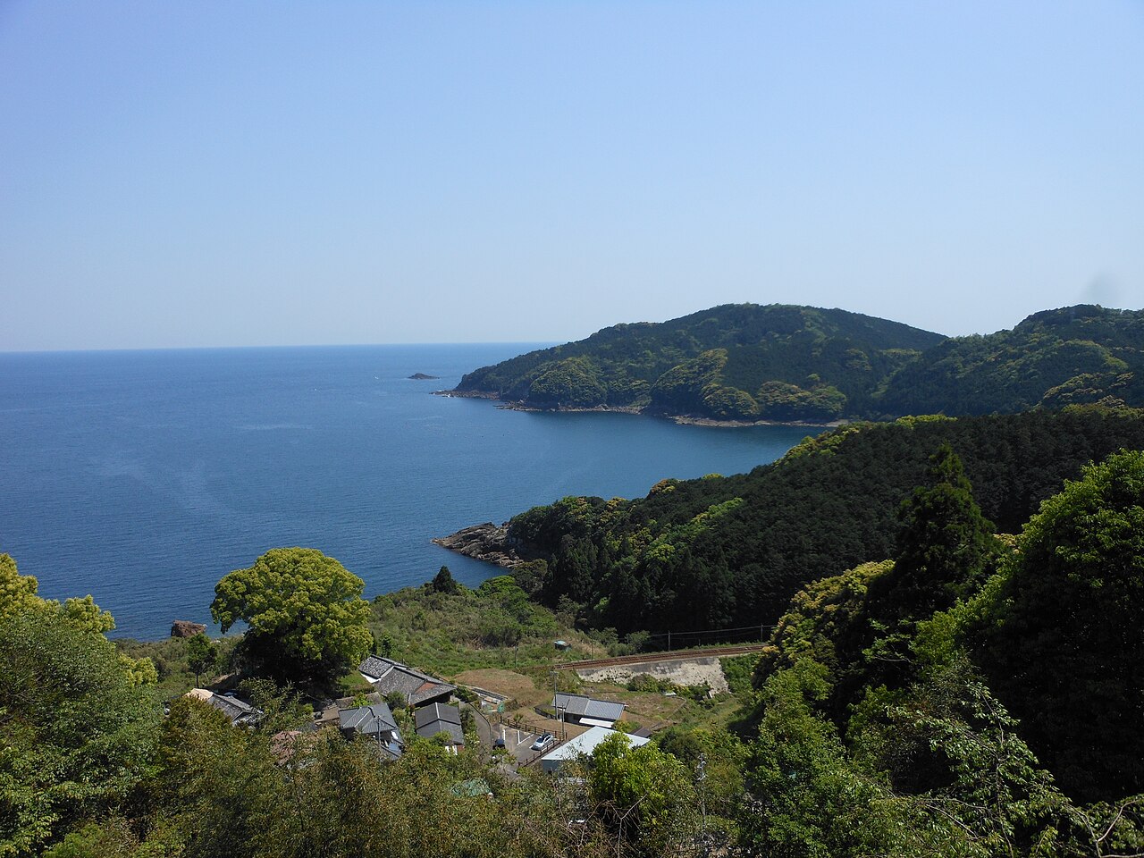 A blue bay and wooded headlands seen from a hillside above a small coastal settlement of tiled roofs