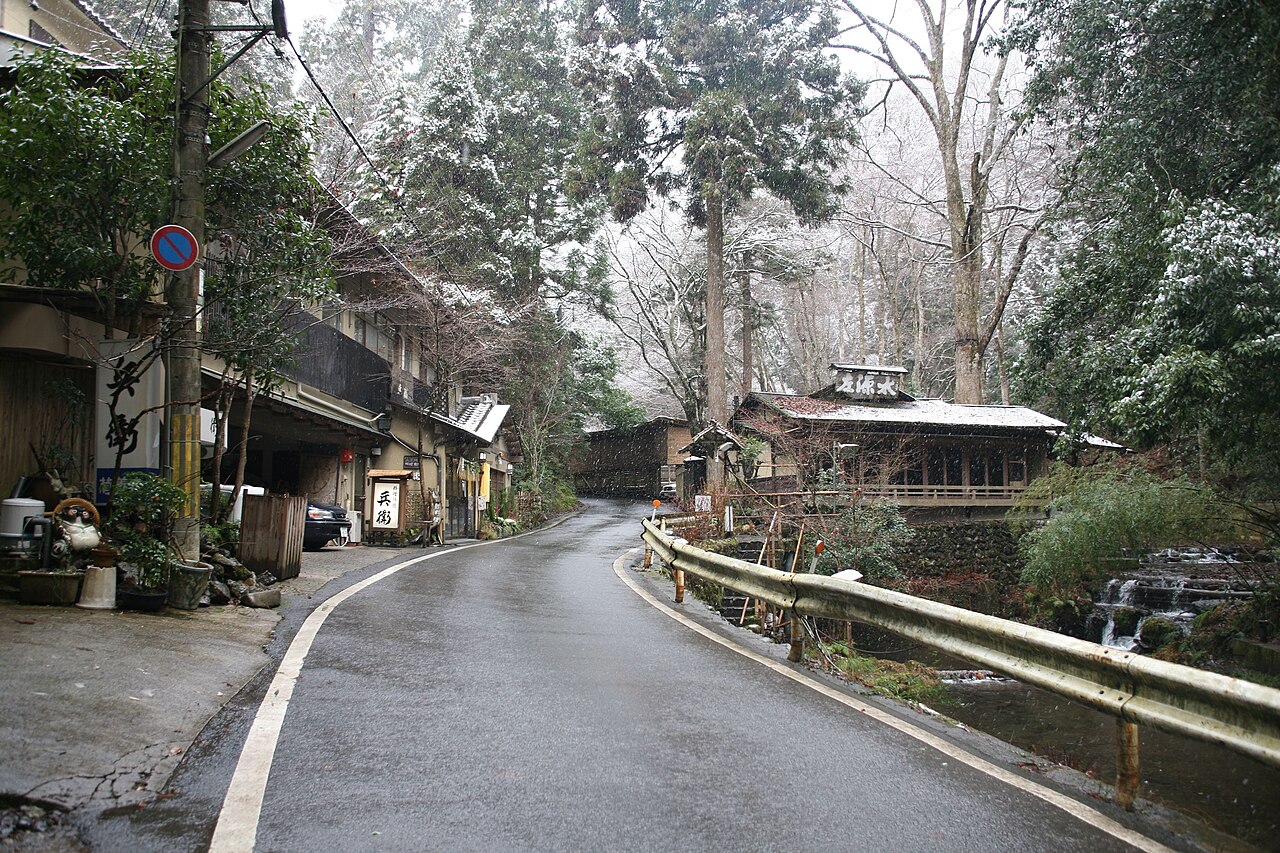 A quiet road curving along a river in a mountain village, with wooden houses and trees dusted with snow on both sides.