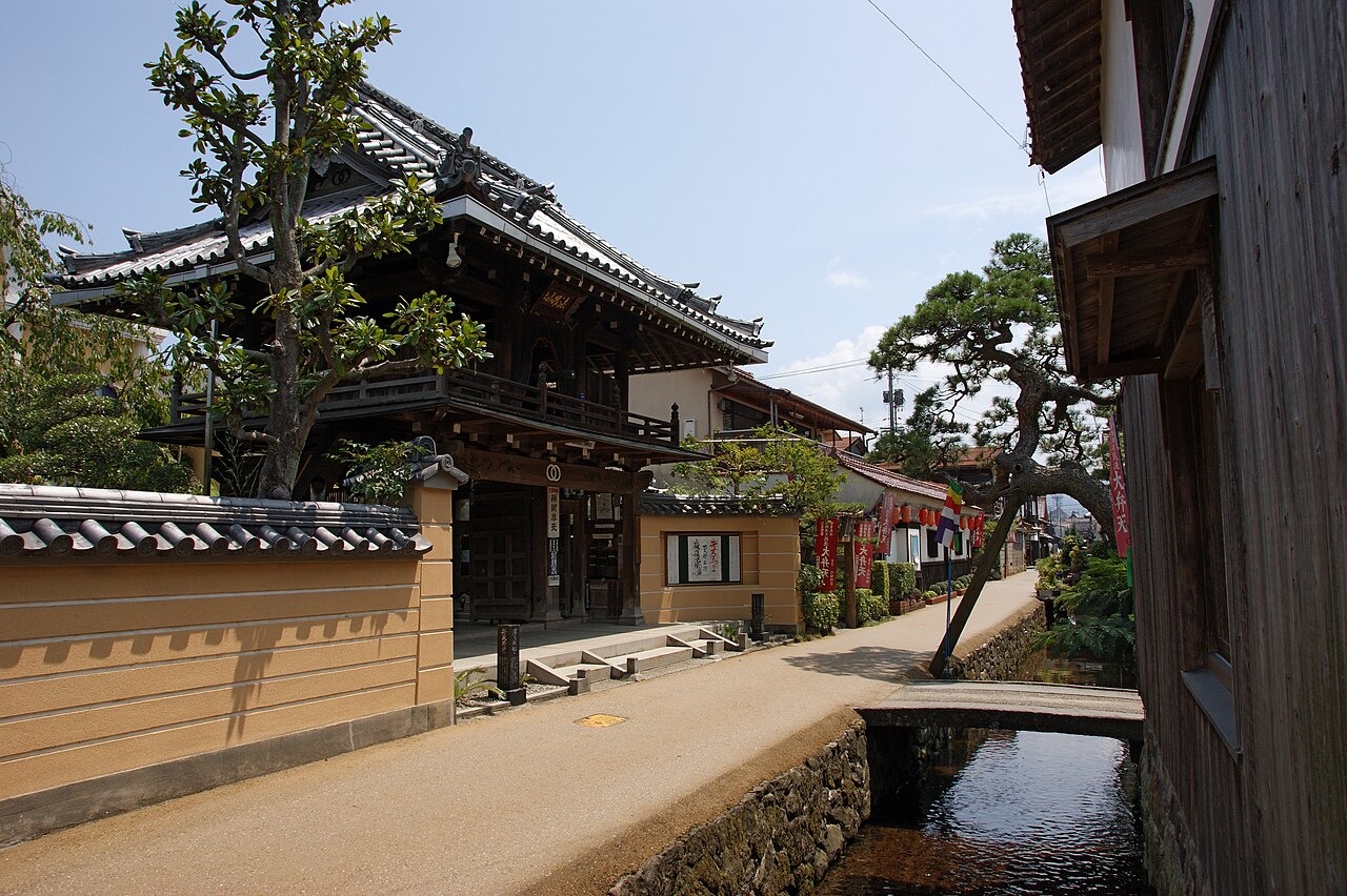 A tiled temple gate with a wooden upper floor beside a narrow stone-lined canal and a small footbridge in Kurayoshi, on a sunny day.