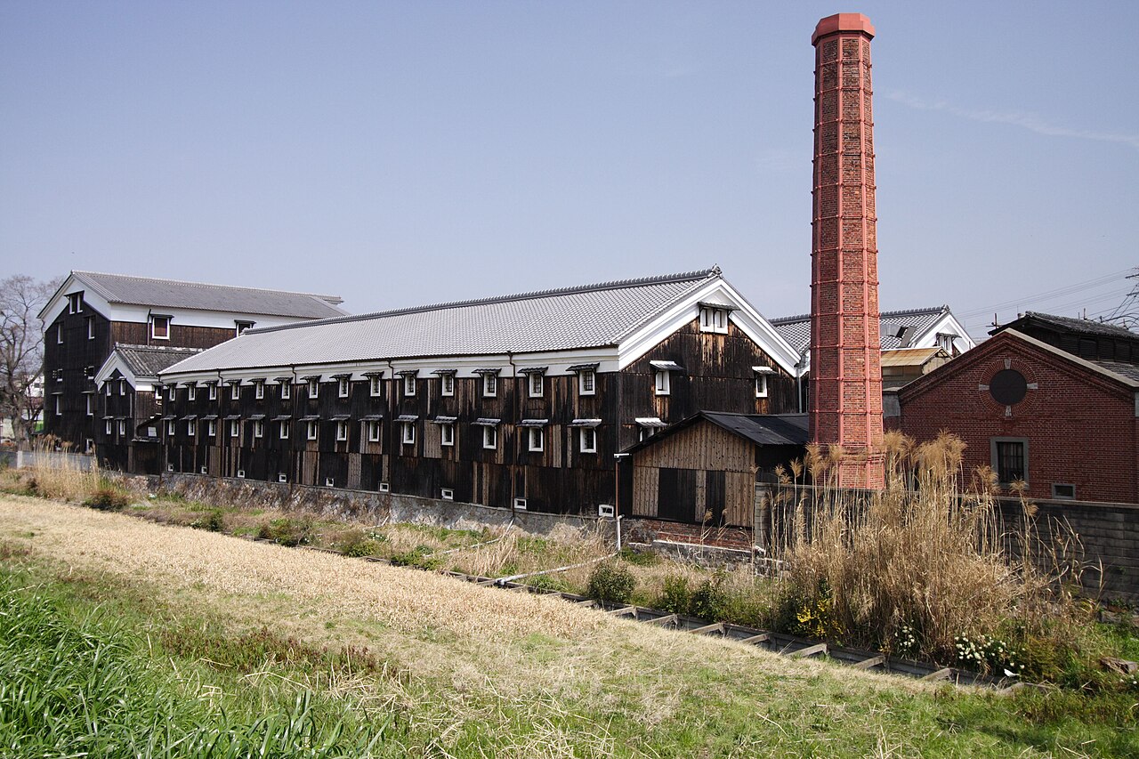 A long dark-timbered sake brewery storehouse with small white-framed windows and a grey tiled roof stands beside a tall red-brick chimney and a brick building, seen across a grassy bank under a pale blue sky.