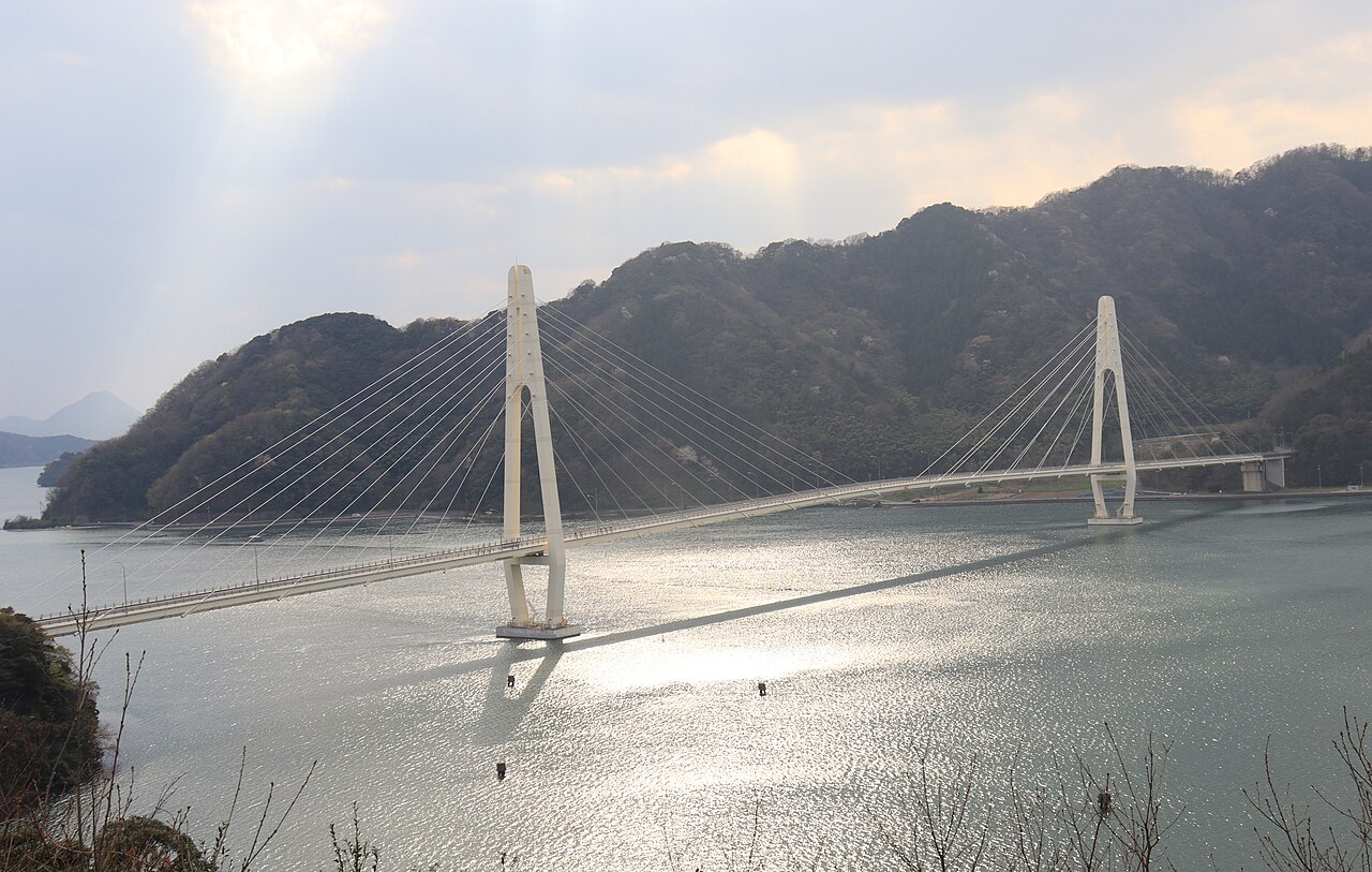 A white cable-stayed bridge with two tall pylons spans a calm bay under a hazy sky with sunbeams, with wooded hills behind and sunlit water in front