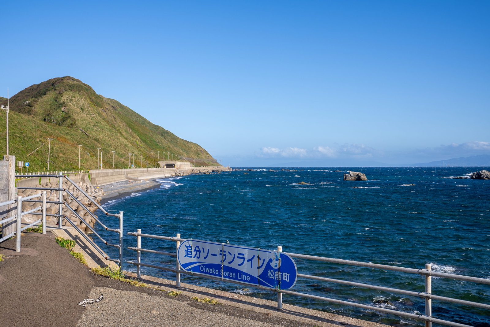 A seaside road with a guardrail along a bright blue sea, with a green headland and a small beach beyond.