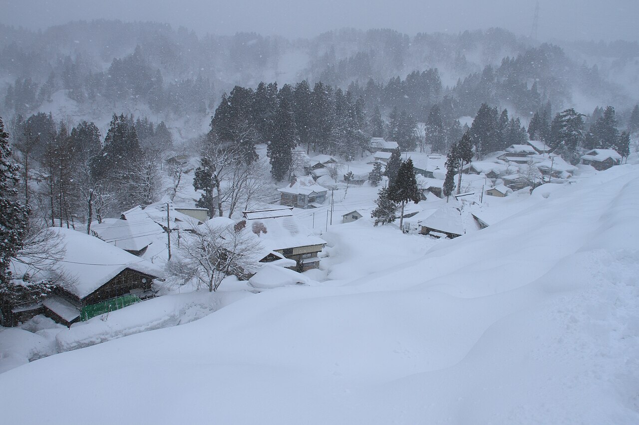 A snow-covered mountain village of low roofs and bare trees under a grey winter sky