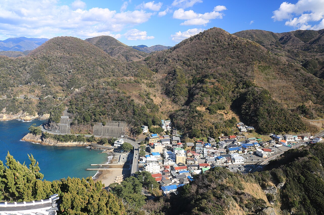 A small fishing village of low houses and a curving coastal road between steep wooded hills, with a blue bay and a sandy cove at the left, seen from above