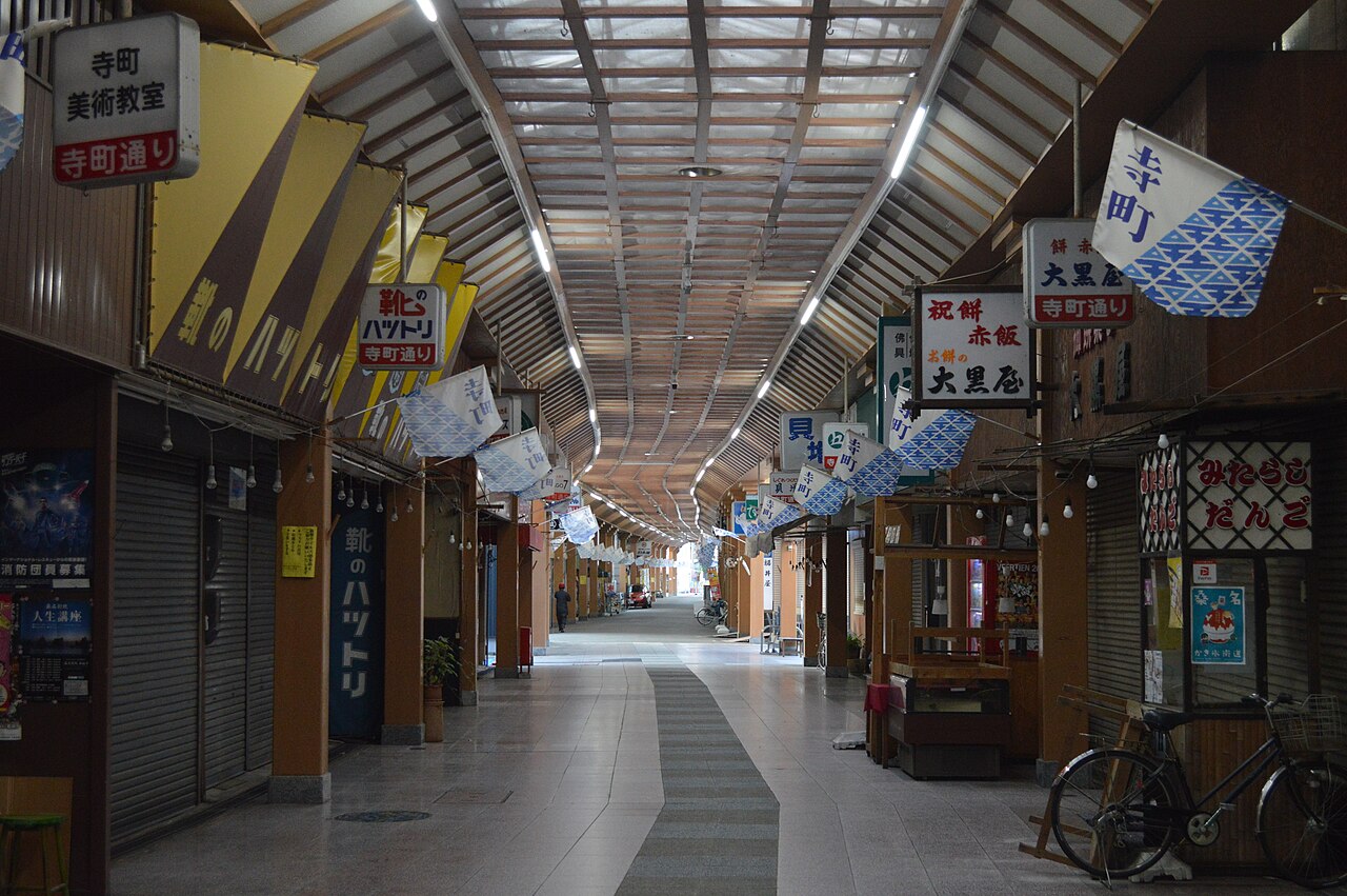 A covered shopping arcade with a wooden-ribbed glass roof, hanging blue and white pennants, shuttered shopfronts, signboards and a parked bicycle, leading down a tiled walkway into the distance.