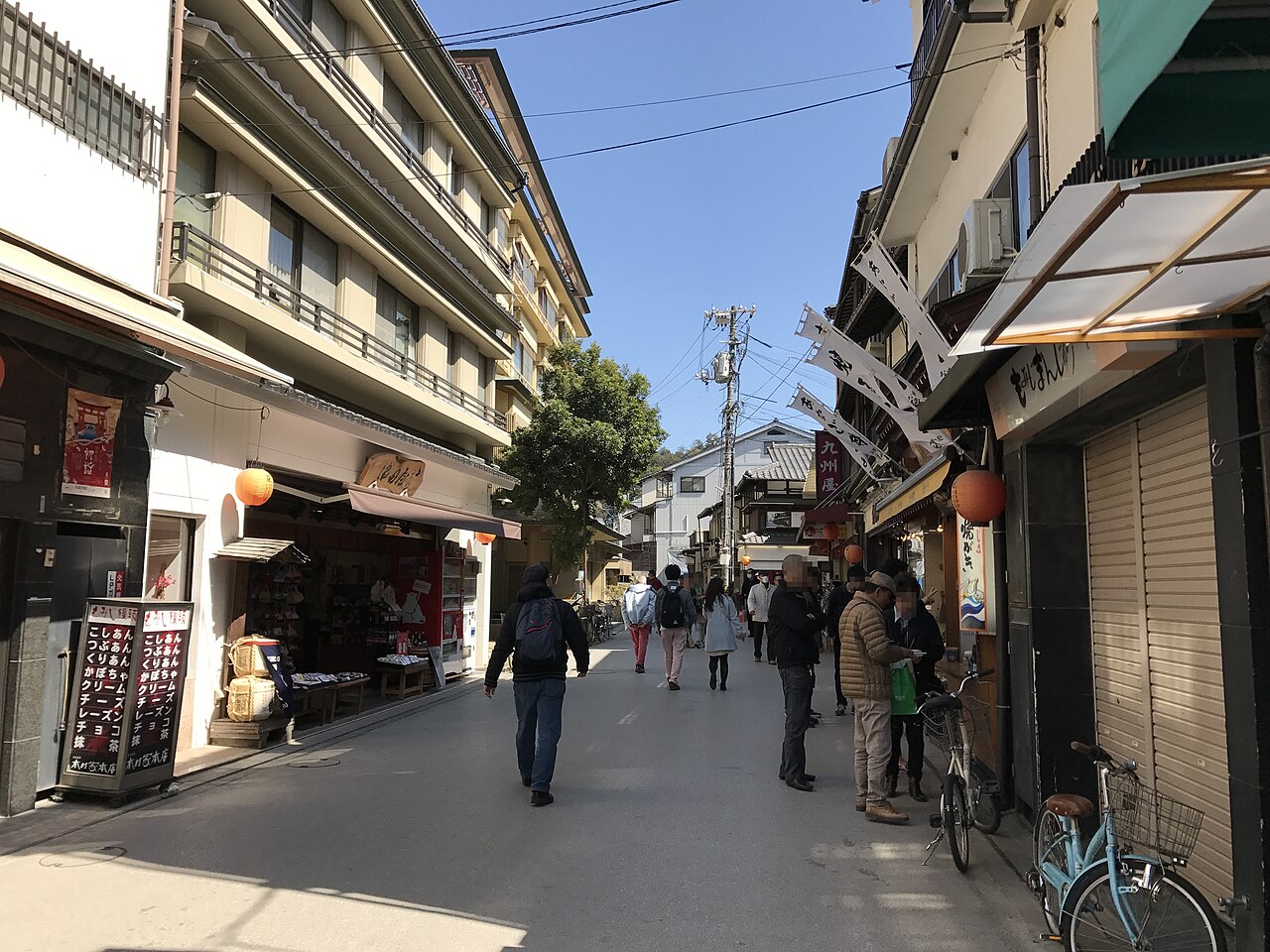 A quiet sunlit street on Miyajima lined with old multi-storey buildings and small shops, with a few visitors walking and a bicycle parked at the roadside.