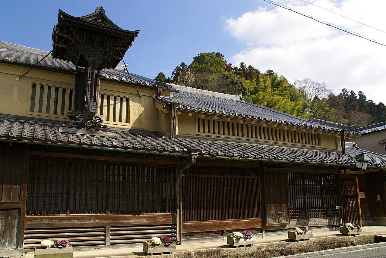 A long traditional merchant house with latticed windows, tiled eaves and a small carved bell-tower roof on a street in Matsuyama, Uda.