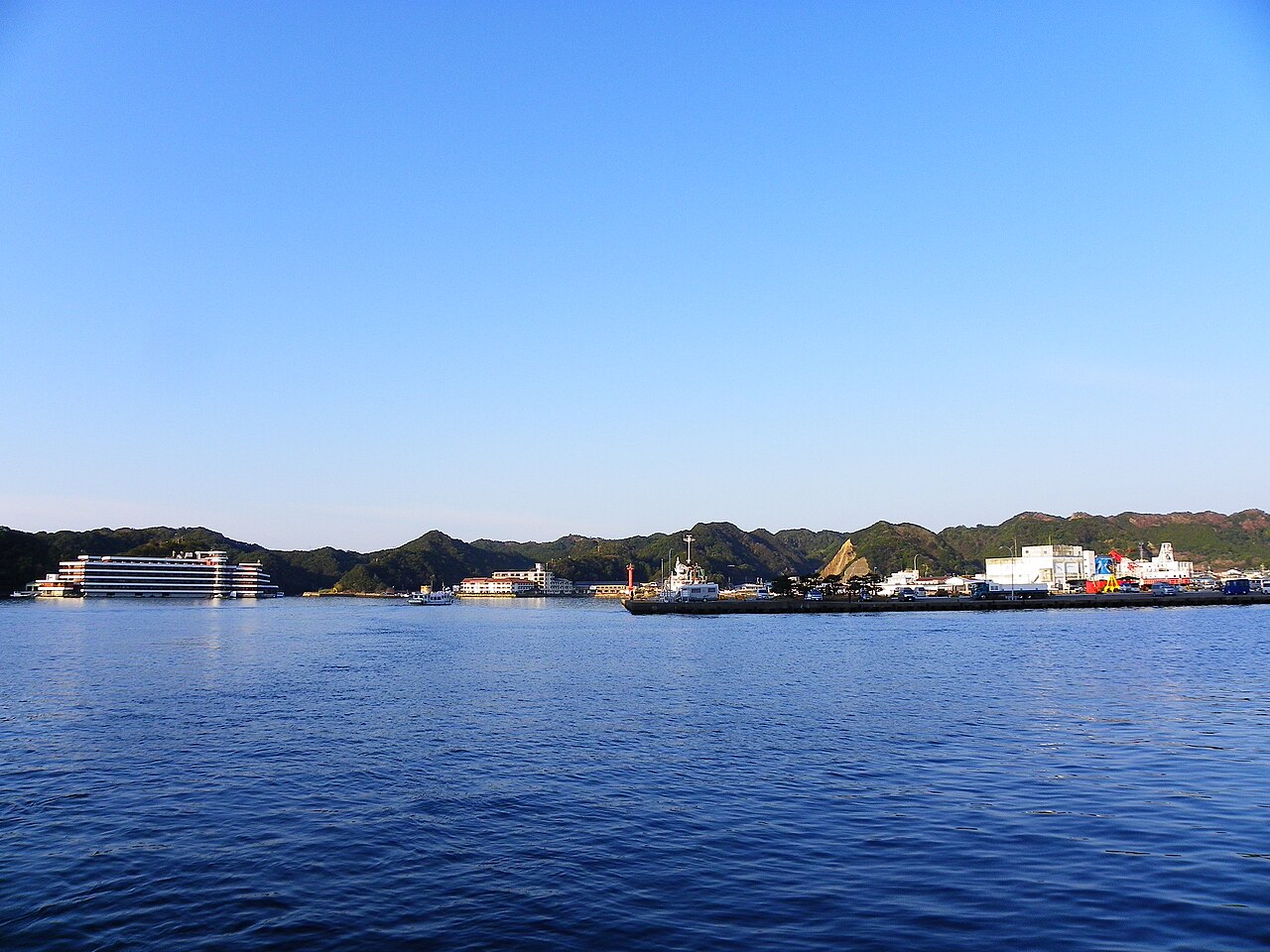 Katsuura Port at calm morning light, with a low waterfront hotel, small pine-covered rocky islets, and moored boats reflected in still blue water, mountains rising behind the harbor.