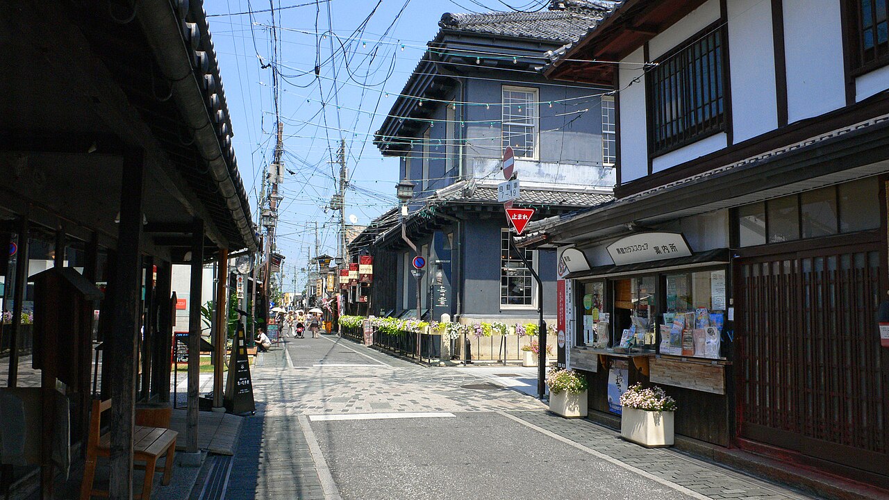 A sunlit lane in Nagahama's old merchant quarter, lined with traditional wooden shopfronts and a grey Western-style building, with flower planters along the roadside.