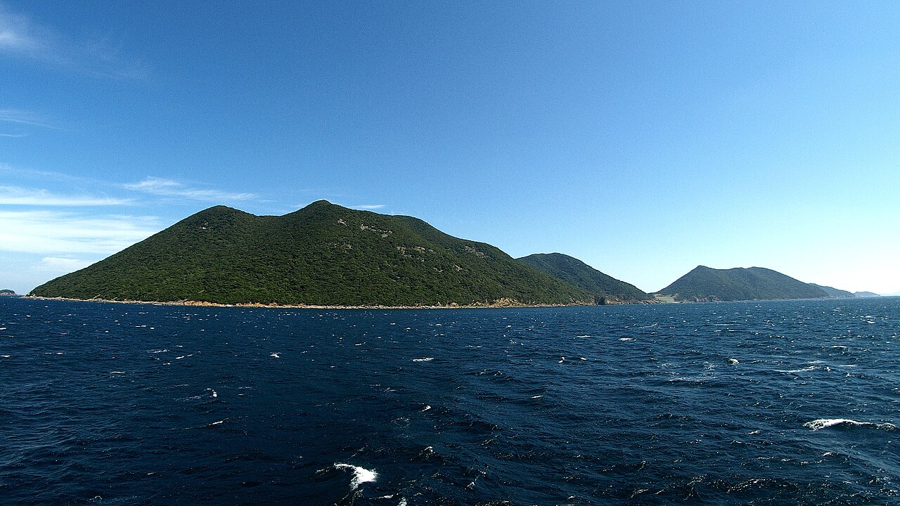 A forested green island with a rounded summit rises from deep blue open water under a clear sky, with another low island beyond to the right.