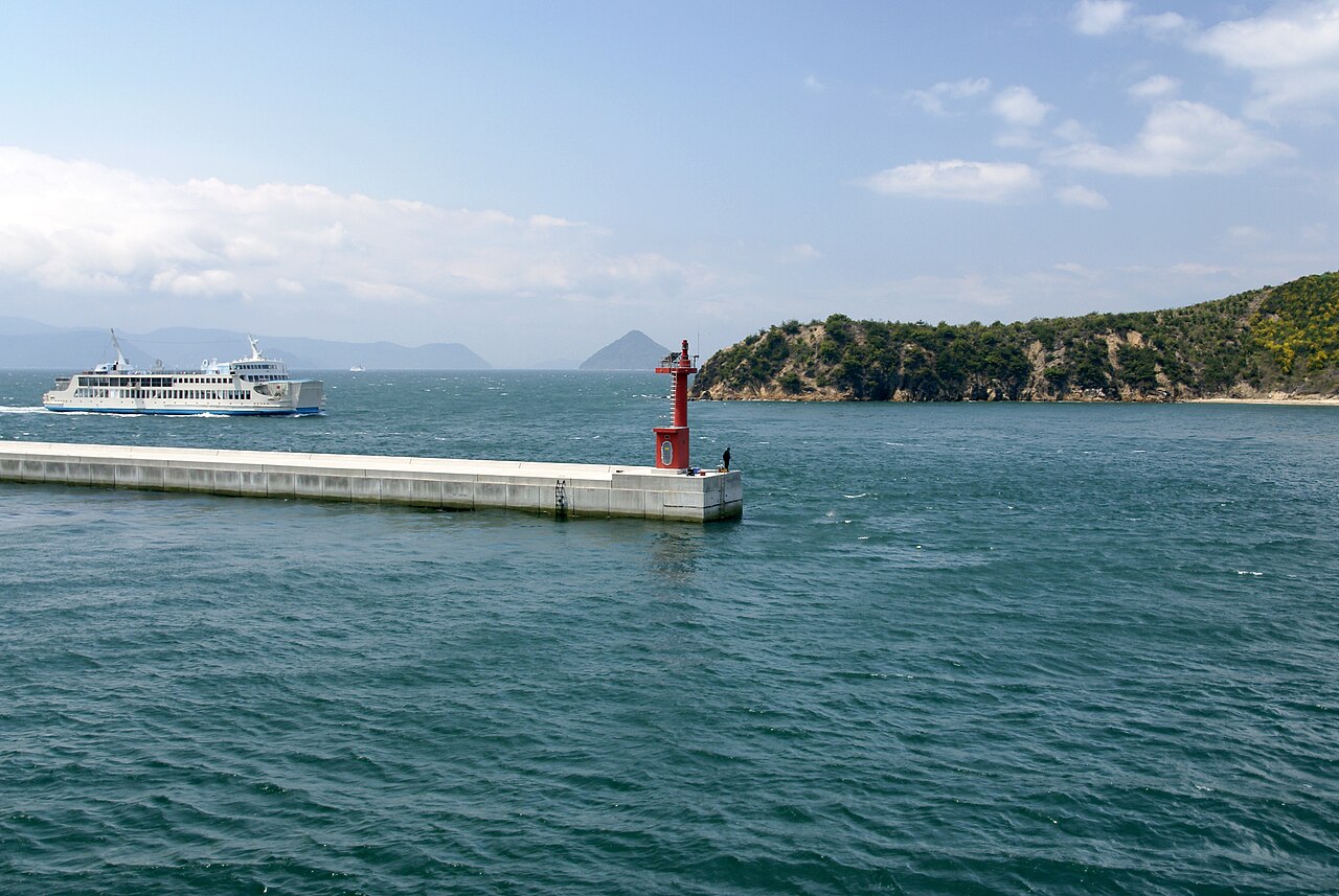 Calm harbour water at Miyanoura on Naoshima with a breakwater, small red lighthouse and a ferry in the distance
