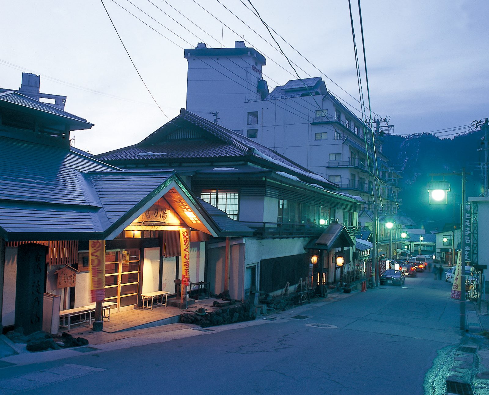 Naruko Onsen main street at dusk, with the lit entrance of a wooden public bathhouse on the left and inn buildings rising behind.