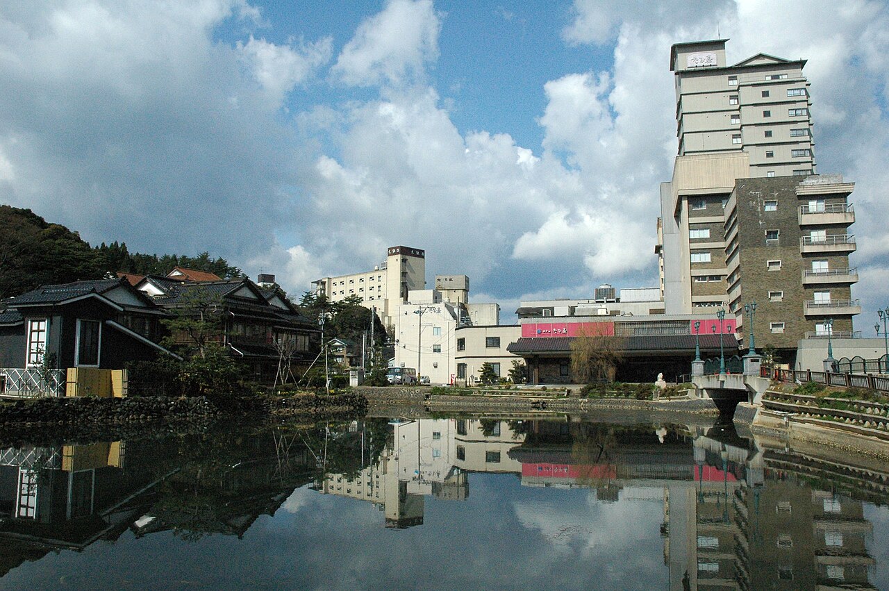 Hot-spring town buildings and old wooden inns stand beside a still pond that mirrors them and a cloudy sky, with a tall hotel at the right